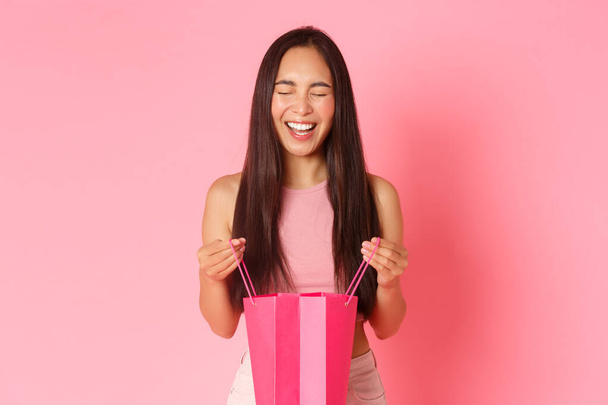 Piękno, moda i koncepcja stylu życia. Zbliżenie podekscytowany i zdumiony, zaskoczony dziewczyna otworzyć prezent, patrząc w torbie na zakupy z radosnym uśmiechem optymistycznym, stojąc różowe tło - Zdjęcie, obraz