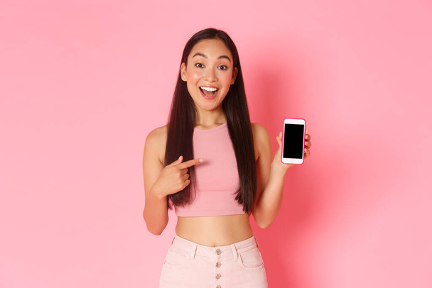 Technology, communication and online lifestyle concept. Amazed happy asian girl in trendy clothes found something online, pointing finger at smartphone screen, pink background - Photo, Image