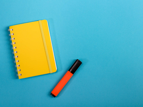 bloc de notas y bolígrafo Un bloc de notas amarillo y un marcador naranja se encuentran a la izquierda sobre un fondo azul con espacio para texto a la derecha, una vista superior de primer plano. - Foto, imagen