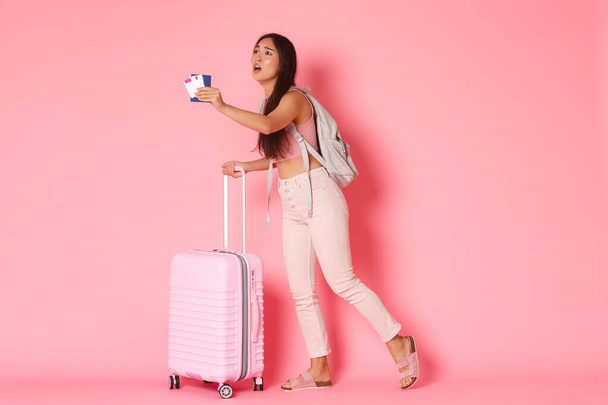 Travelling, holidays and vacation concept. Upset gloomy asian girl tourised missed her flight, looking with regret and dispair at departure gates in airport, standing with suitcase and passport - Photo, Image