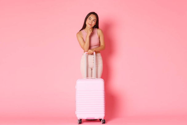 Viaggiare, vacanze e concetto di vacanza. Ragazza bella asiatica premurosa e curiosa con valigia, guardando a sinistra incuriosita, pensando a dove viaggiare, tour di raccolta turistica in agenzia, sfondo rosa - Foto, immagini