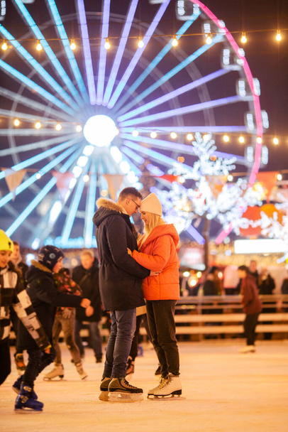 Man and woman in love in embrace on ice arena against of Ferris wheel in evening of Christmas. Couple at city skating rink, romance and glowing lanterns, light bulbs and garlands festive atmosphere. - Photo, Image