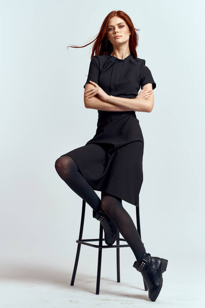 Yüksek sandalyede oturan bir kadın. Uzun boylu, siyah elbiseli, kızıl saçlı, model botlu. - Fotoğraf, Görsel