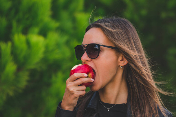 Mulher bonita jovem morena vegan em óculos de sol pretos comendo maçã vermelha saborosa no fundo árvores verdes. Morde a maçã. Conceito de estilo de vida saudável. - Foto, Imagem