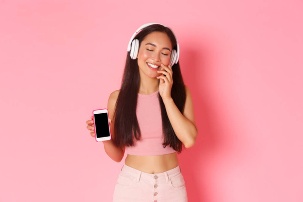 Technology, communication and online lifestyle concept. Lovely, dreamy asian girl in headphones, close eyes and touching cheek silly, daydreaming during listening music, showing mobile screen - 写真・画像