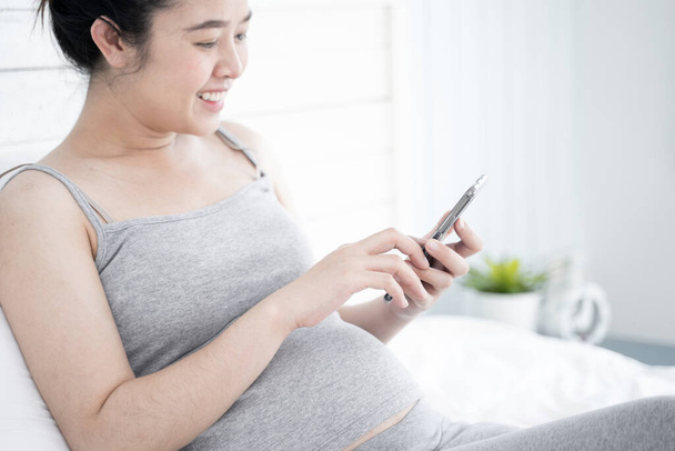 Beautiful pregnant woman using mobile phone in her bedroom. - Фото, изображение