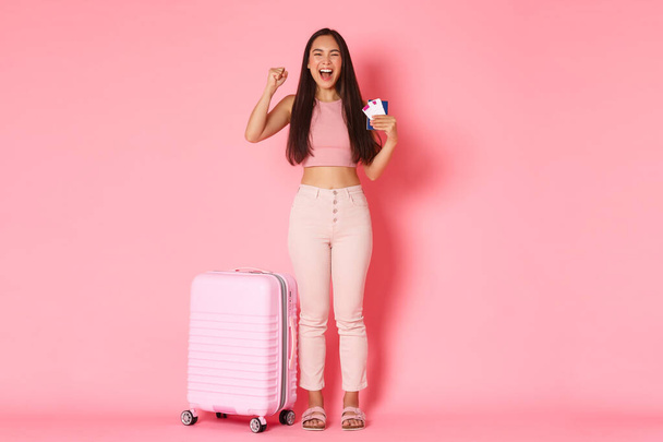 Travelling, holidays and vacation concept. Full-length of cheerful, rejoicing asian girl tourist finally going abroad after quarantine, pandemic restrictions, raising hand up, showing flight tickets - Photo, Image
