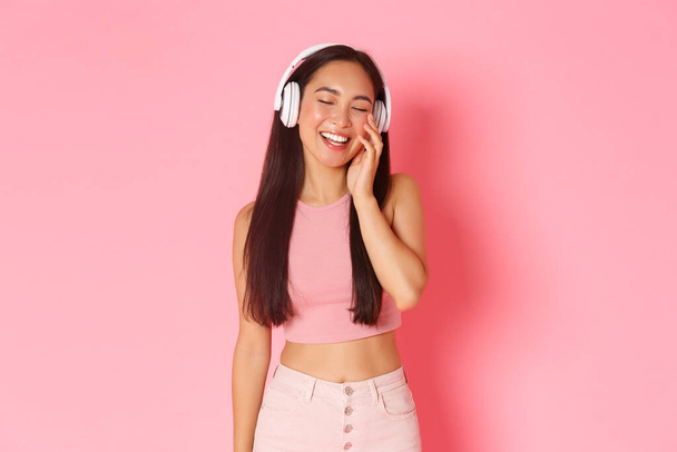 Portrét potěšené a šťastné, usmívající se atraktivní asijské dívky v bezdrátových sluchátkách, poslouchající hudba nebo legrační podcast, vesele zavřené oči, stojící nad růžovým pozadím - Fotografie, Obrázek