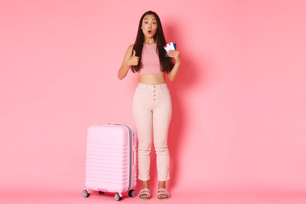 Utazás, nyaralás és nyaralás koncepció. Nyári ruhában, bőrönd, útlevél és repülőjegy közelében álló, izgatott ázsiai turista lány teljes hossza, hüvelykujj-felhúzás, légitársaságot ajánló - Fotó, kép