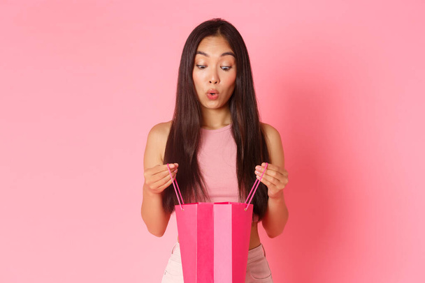 Beauty, fashion and lifestyle concept. Close-up of excited and amazed, surprised girl open up present, looking in shopping bag with impressed and wondered expression, pink background - Foto, immagini