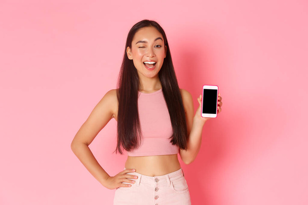 Technologie, Kommunikation und Online-Lifestyle-Konzept. Porträt kokett zwinkernden asiatischen Mädchen ermutigen Download-Anwendung, Besuche Podcast oder Internet-Shop, zeigt Smartphone-Bildschirm - Foto, Bild