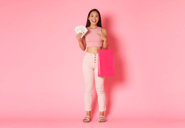 В длину богатая привлекательная девушка в летней одежде, наслаждаясь покупками, имеет деньги, указывая пальцем на наличные деньги, которые тратятся на магазины и магазины, стоящие на розовом фоне - Фото, изображение