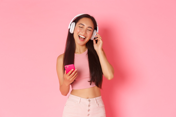Retrato en la cintura de la atractiva chica asiática con estilo cerrar los ojos y cantar a lo largo de la canción mientras escucha música en los auriculares, juego de karaoke en el teléfono móvil, de pie sobre el fondo rosa - Foto, Imagen