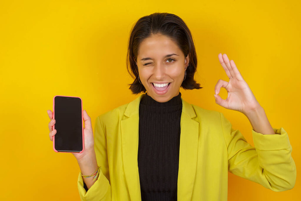 hermosa joven con teléfono inteligente posando contra la pared amarilla, stusio disparo - Foto, imagen