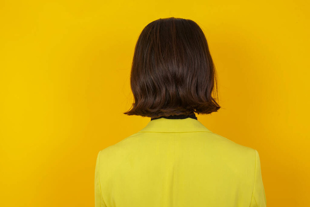 Het achteraanzicht van een mooie jonge vrouw die tegen een gele muur staat. Studio Shoot. - Foto, afbeelding