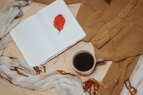 an open daily diary on empty pages, Cup of hot coffee, beige cardigan with buttons,garland laid out in a spiral lie on a wooden light table. Red leaves lie on a sweater, Notepad and table. Top view. - Foto, afbeelding