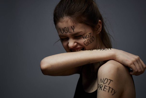 beleidigte Frau mit wütenden Schriftzügen auf ihrem Körper berührt sich mit den Händen auf grauem Grund  - Foto, Bild
