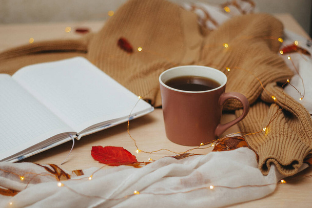 an open daily diary on empty pages, Cup of hot coffee, beige cardigan with buttons,garland laid out in a spiral lie on a wooden light table. Red leaves lie on a sweater, Notepad and table. Side view. - Fotó, kép