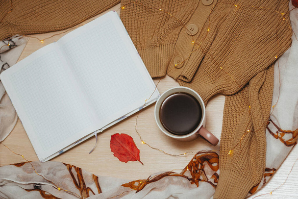 an open daily diary on empty pages, Cup of hot coffee, beige cardigan with buttons,garland laid out in a spiral lie on a wooden light table. Red leaves lie on a sweater, Notepad and table. Top view. - Foto, Imagem