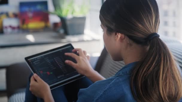 Молодая взрослая женщина лежит на диване в гостиной дома, девушка торгует онлайн с планшетным компьютером, подключенным к фондовым рынкам. Люди, финансы, инвестиции. Вид через плечо - Кадры, видео