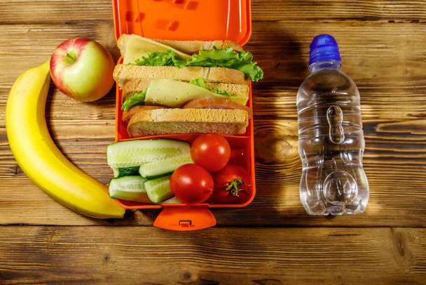 Botella de agua, manzana, plátano y lonchera con sándwiches y verduras frescas sobre una mesa de madera. Vista superior - Foto, Imagen