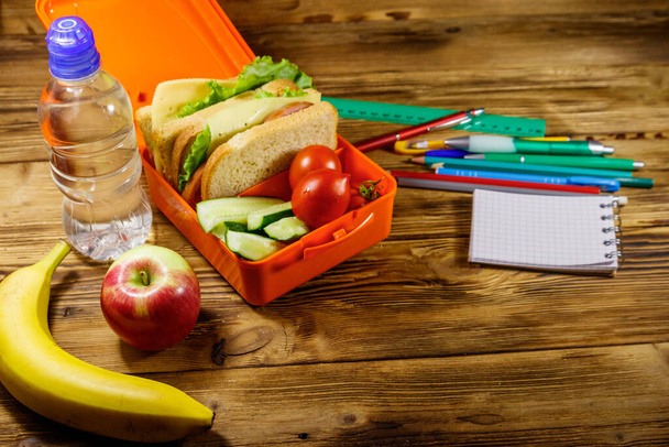 Back to school concept. School supplies, bottle of water, apple, banana and lunch box with sandwiches and fresh vegetables on a wooden desk - Photo, image