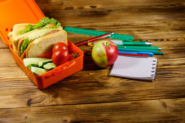 Вернемся к школьной концепции. Школьные принадлежности, яблоко и обеденная коробка со сэндвичами и свежими овощами на деревянном столе - Фото, изображение