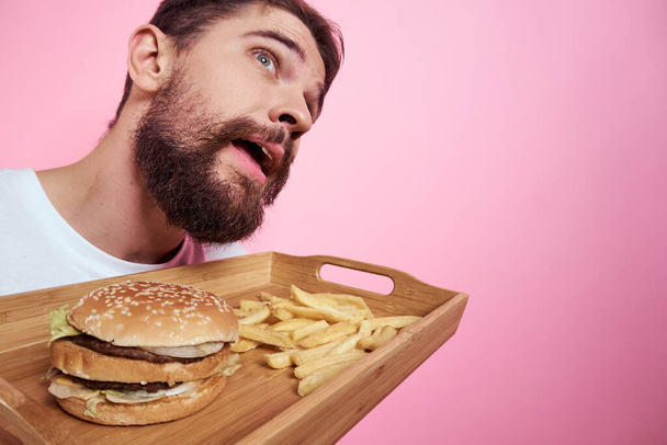 Homem com uma bandeja de comida na mão hambúrguer batatas fritas e fast food caloria fundo rosa retrato close-up - Foto, Imagem
