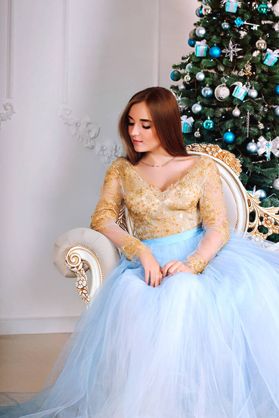 Young and beautiful woman in long party dress sitting near Christmas tree in studio - Fotografie, Obrázek