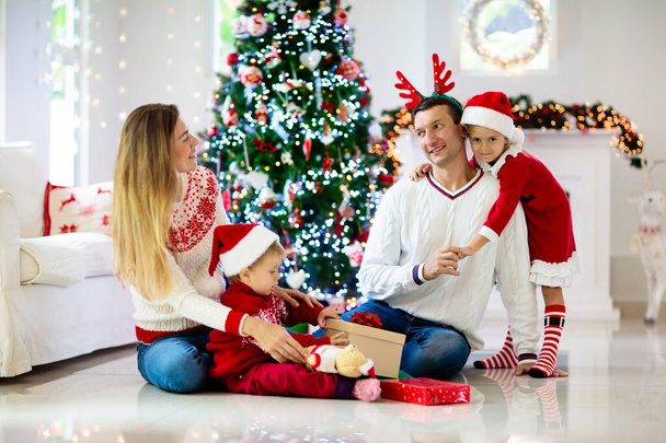Perhe viettää joulua. Vanhemmat ja lapset koristella joulukuusi ja avaa lahjoja. Lapsi, jolla on laatikko. Lapsi avaamassa lahjoja. Kodin sisustus talvilomalle. Juhlava sisustettu olohuone. - Valokuva, kuva