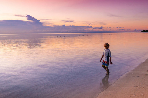 Ребенок играет на пляже океана. Мальчик прыгает по волнам на закате. Морской отдых для семьи с детьми. Маленький мальчик на тропическом пляже экзотического острова во время летнего отдыха. Вид на закат Мбаппе и водные развлечения. - Фото, изображение