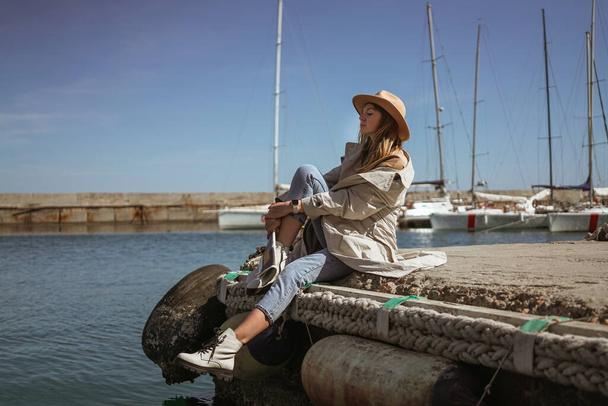 Egy lány fehér cipőben és kalapban ül a mólón egy jachtklubban, újságot olvas és pihen. új kollekció lábbeli ősz tél 20 / 21 divat fotó - Fotó, kép