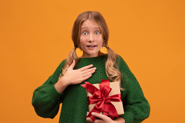 Image of charming blonde girl 12-14 years old in warm green sweater holding present box with red bow. Studio shot, yellow background, isolated. New Year Women's Day birthday holiday concept - Foto, Imagem