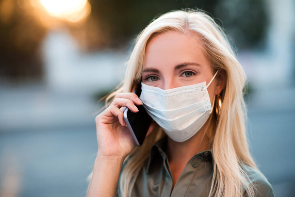 Young blonde woman 24-26 year old wearing medical protective mask talking on phone walking in street outdoors close up. Looking at camera. Coronavirus pandemic. Stay at home. Healthcare. - Photo, Image