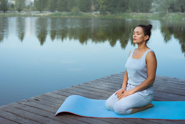 Молода жінка сидить на килимку біля озера, тримаючи релігійний розарій закритими очима. Медитація в природі
. - Фото, зображення