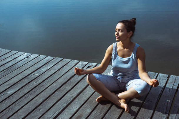 The young woman on a wooden pier sits in a lotus position with her head turned to the side with her eyes closed. Meditation, yoga in nature. - 写真・画像