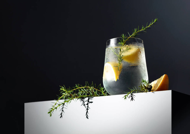 Alcohol drink (gin tonic cocktail) with lemon, juniper branch, and ice on a dark reflective background, copy space. Iced cocktail drink with lemon.  - Fotó, kép