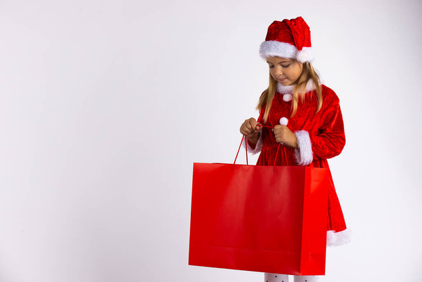Child holding and looks in red bag after shopping. Christmas girl on winter sale with gift, isolated on white background - Foto, immagini