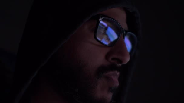 Asijský muž s kapucí a reflexí počítačových obrazovek v brýlích v tmavé místnosti. Uzamčeno, boční pohled - Záběry, video