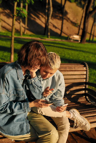 Two young women holding their smartphone, sitting on the bench in the park. Lesbian couple using their phones while spending time together outdoors - Fotoğraf, Görsel
