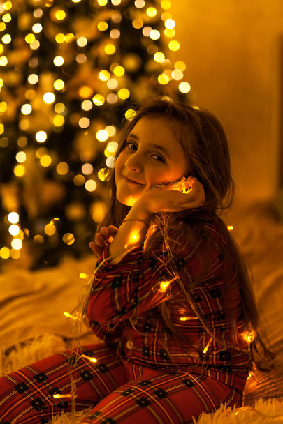 magische mysterieuze meisje kind met een blik met een lichtgevende bloemenslinger, klassieke donkere achtergrond en warm licht. concept van de verwachting van Kerstmis, gezellige warme stijl - Foto, afbeelding