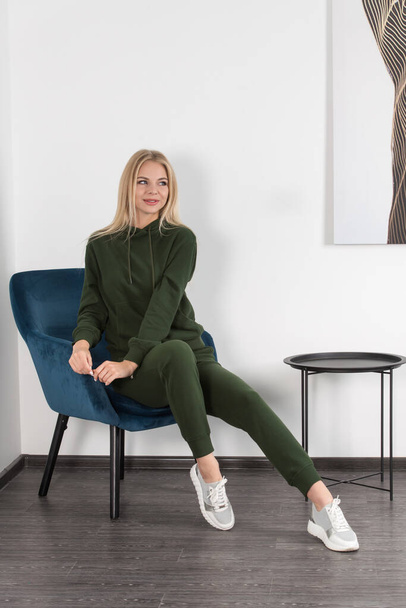 Stijlvolle mooie jonge blonde vrouw in een groen trainingspak poseert in de buurt van een witte muur in de kamer. Aantrekkelijk meisje model poseren in een blauwe stoel. - Foto, afbeelding