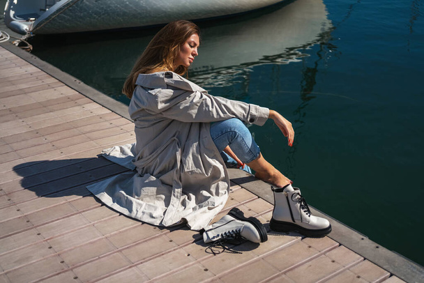 όμορφη γυναίκα κάθεται ξυπόλητη στην προβλήτα φορώντας παλτό και τζιν. λευκά παπούτσια νέα συλλογή παπουτσιών φθινόπωρο χειμώνα 20 / 21 - Φωτογραφία, εικόνα