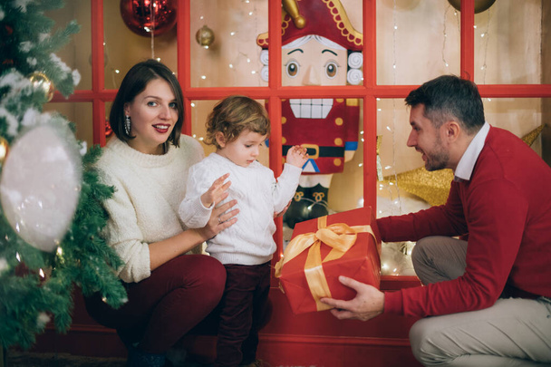Семья с ребенком возле новогодней елки в рождественских украшениях. Малыш и мама с папой веселятся и играют на фоне гирлянд и праздничных огней. - Фото, изображение