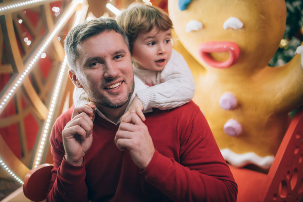 お父さん、小さな男の子とクリスマスの装飾で新年の木の近くに巨大なクッキー。ガーランド、装飾品やお祝いのライトの背景に面白い赤ちゃんと彼の父. - 写真・画像
