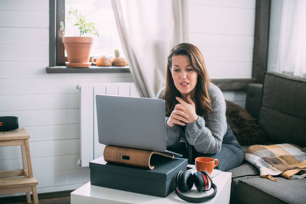 Μια γυναίκα κάθεται στο σαλόνι με ένα φορητό υπολογιστή, κοιτάζει την οθόνη, κρατά σημειώσεις, μαθαίνει μια ξένη γλώσσα στο Διαδίκτυο, διαβάζει ένα online μάθημα κατάρτισης, εκπαίδευση στο Διαδίκτυο, συμβουλεύεται έναν πελάτη σε βίντεο - Φωτογραφία, εικόνα