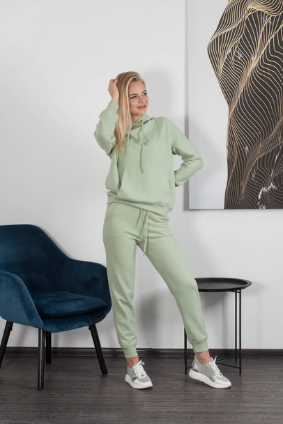 Stylish beautiful young blond woman in a light green tracksuit poses near a white wall in the room. Attractive girl model posing near blue chair. - Photo, image