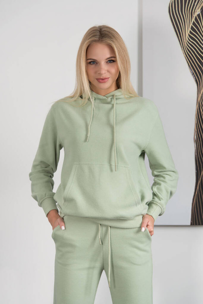 Stylish beautiful young blond woman in a green tracksuit poses near a white wall in the room. - Fotó, kép