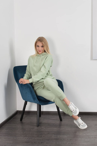 Elegante bella giovane donna bionda in una tuta verde chiaro posa vicino a un muro bianco nella stanza. Attraente ragazza modello in posa su una sedia blu. - Foto, immagini