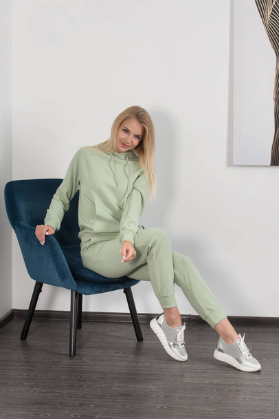 Stylish beautiful young blond woman in a light green tracksuit poses near a white wall in the room. Attractive girl model posing in a blue chair. - Photo, image
